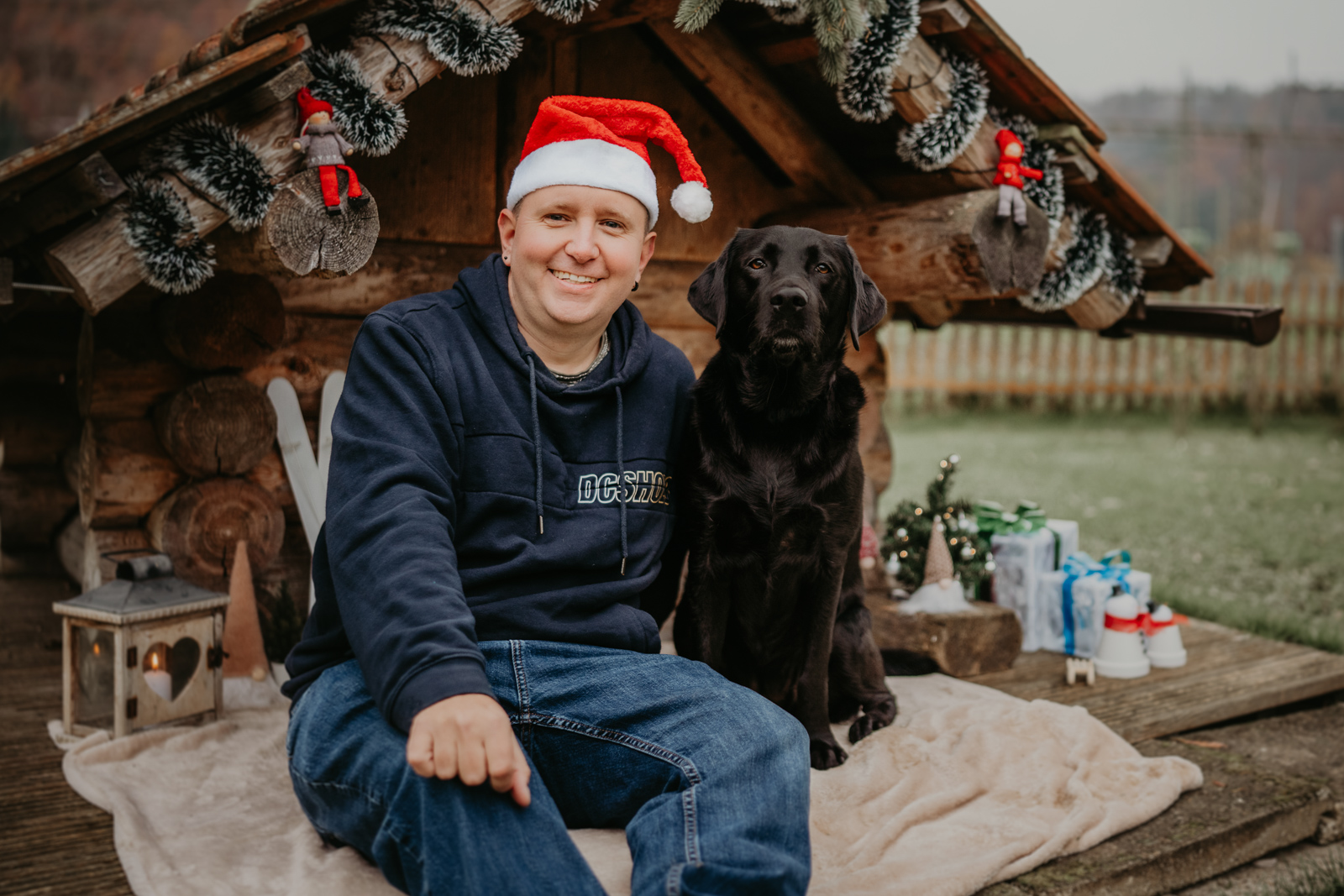 227 - lisamariekurth-hundefotografin-weihnachten-223 Hundefotografie zu Weihnachten: Besitzer und sein Hund vor einer weihnachtlich geschmückten Holzhütte.