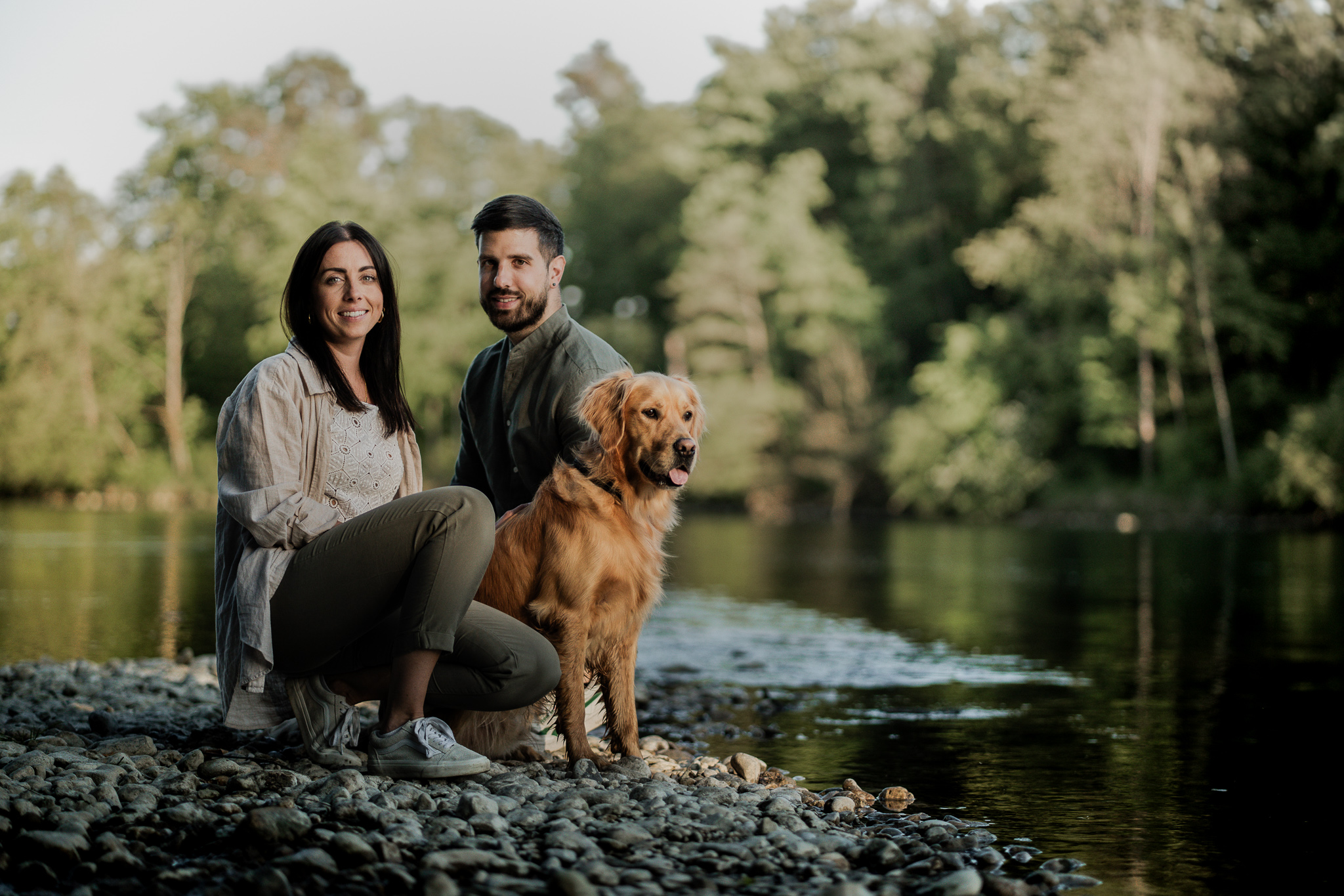 Paar sitzt mit Golden Retriever am Ufer eines ruhigen Flusses inmitten grüner Natur.