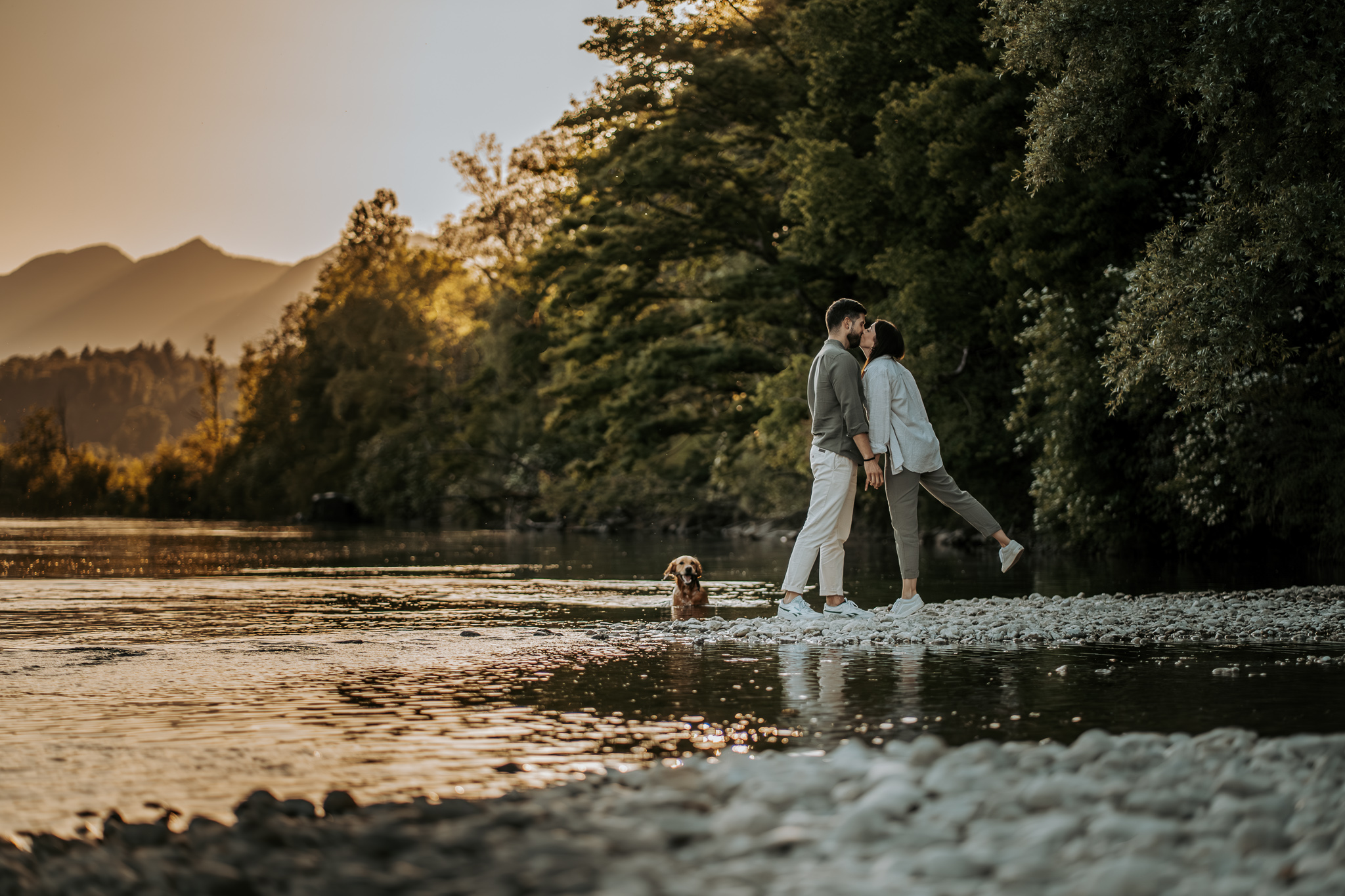 Paar küsst sich auf Kiesbank an einem Fluss bei Sonnenuntergang, während ihr Golden Retriever im Wasser steht.
