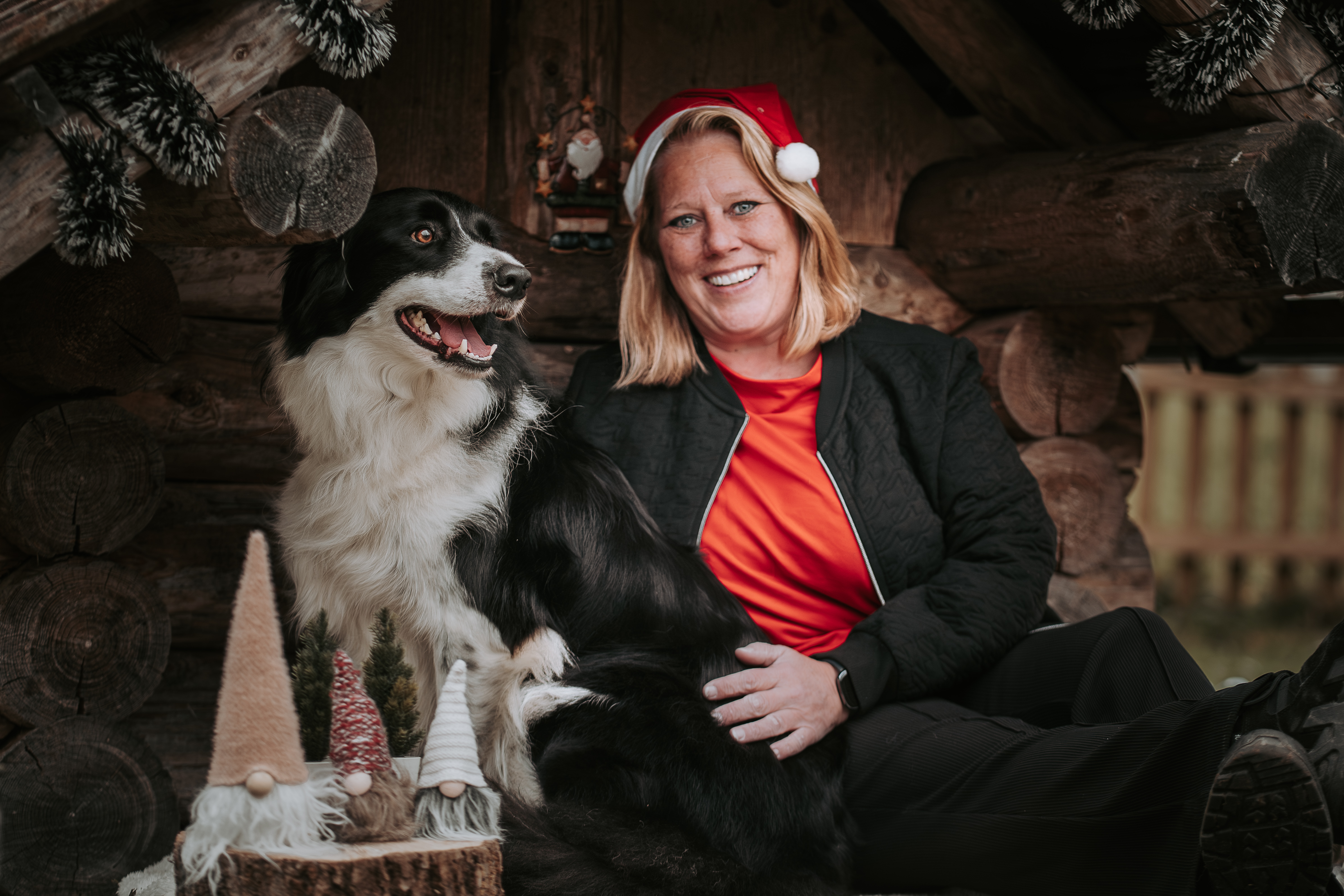 Hund und Halterin sitzen gemeinsam in einer rustikalen Holzhütte, beide lachen, der Hund blickt liebevoll zu seiner Bezugsperson – emotionale Mensch-Hund-Fotografie zu Weihnachten.