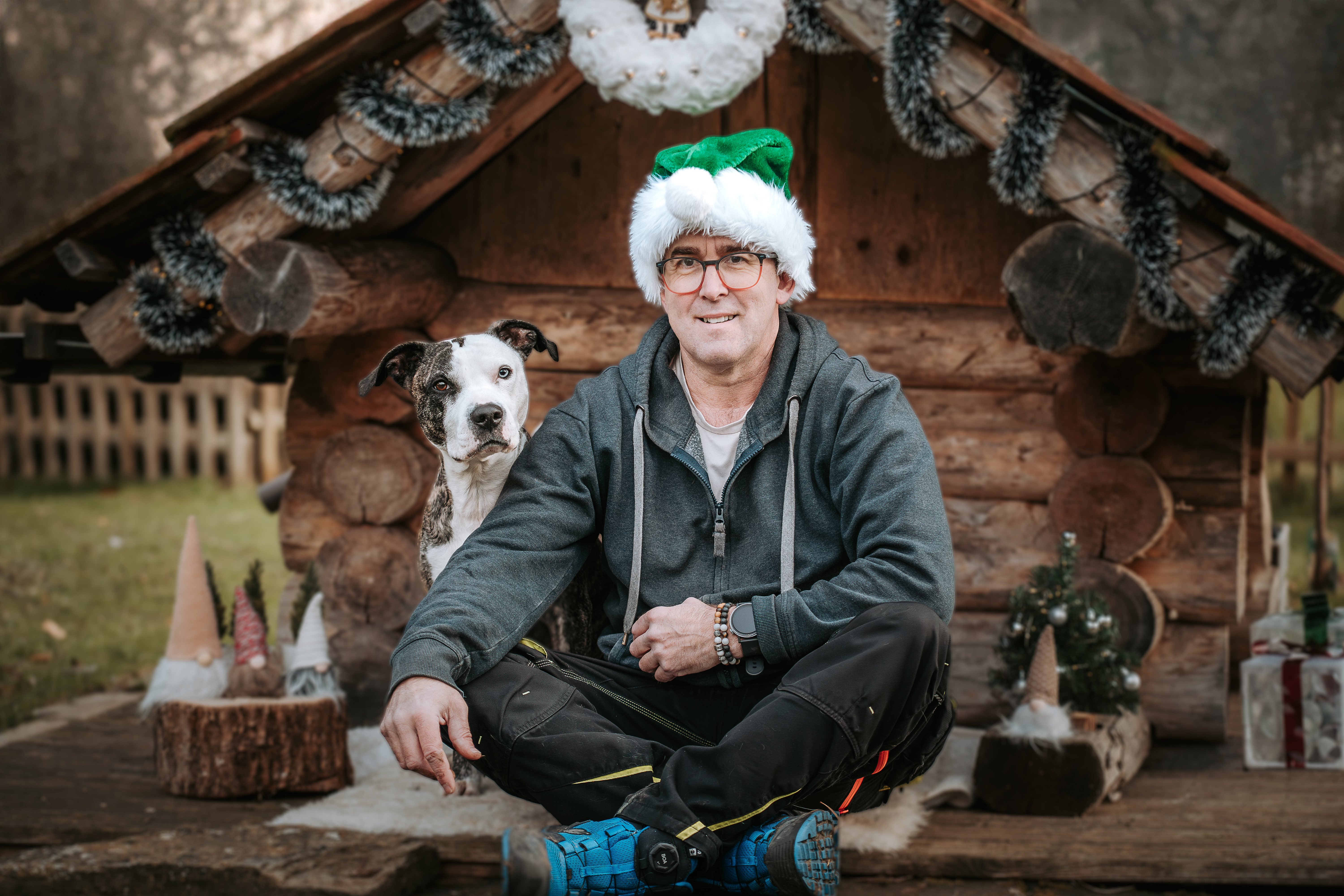 Mann mit Weihnachtsmütze sitzt vor einer rustikalen Holzhütte, sein Hund sitzt ruhig neben ihm, natürliche Weihnachtsdekoration und Outdoor-Umgebung – emotionale Hundefotografie von Lisa-Marie Kurth.